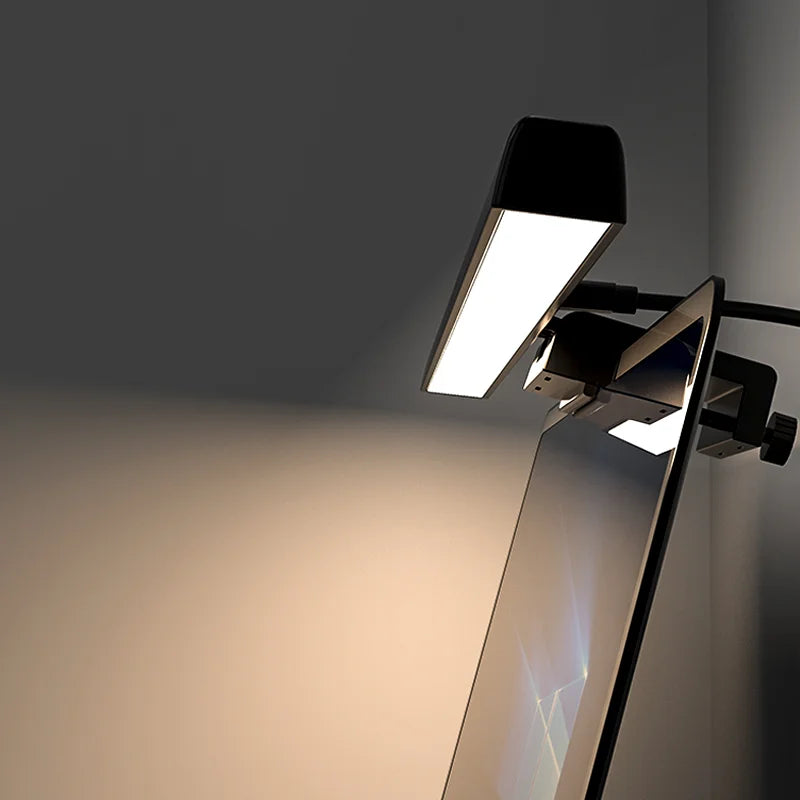 Clamp-on desk lamp with a folding head lit warm white beside a monitor